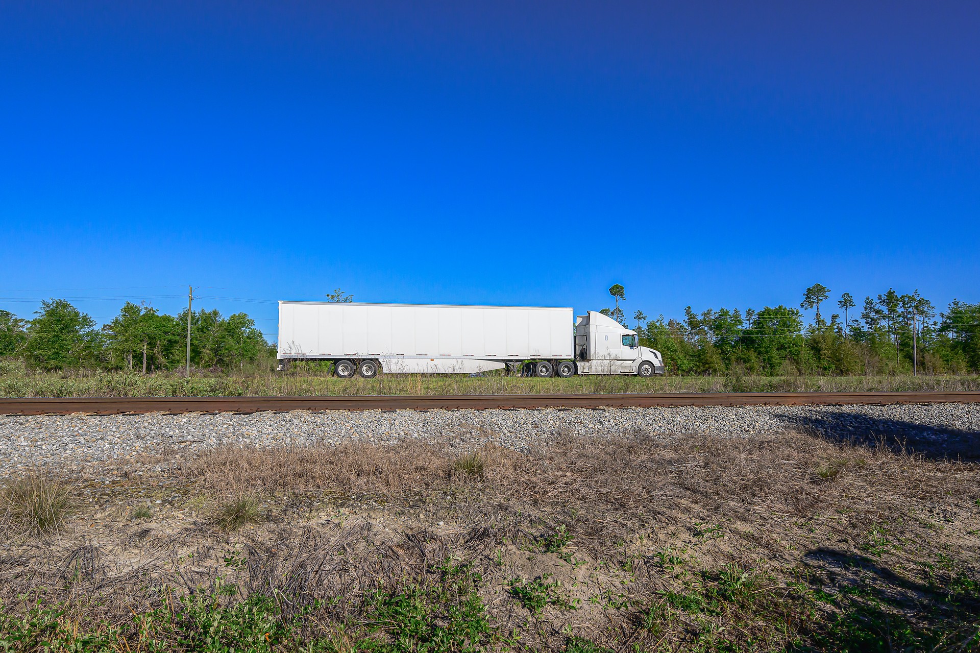 colossal 18-wheeler tractor-trailer traverses the highway, as railroad tracks stretch in the foreground against the backdrop of a brilliant blue sky